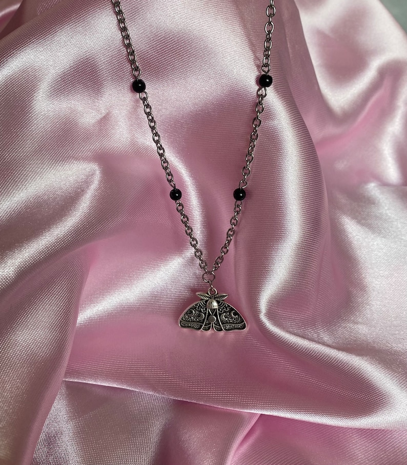 K&ouml;nnte beinhalten: Silberfarbene Halskette mit einem Anh&auml;nger in Form einer Motte. Die Kette hat kleine schwarze Perlen. Der Mottenanh&auml;nger ist mit komplizierten Mustern versehen, vor einem weichen, rosa Satinhintergrund.