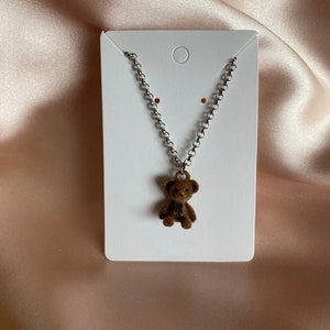 May include: A silver chain necklace with a brown teddy bear charm. The necklace is displayed on a white card.