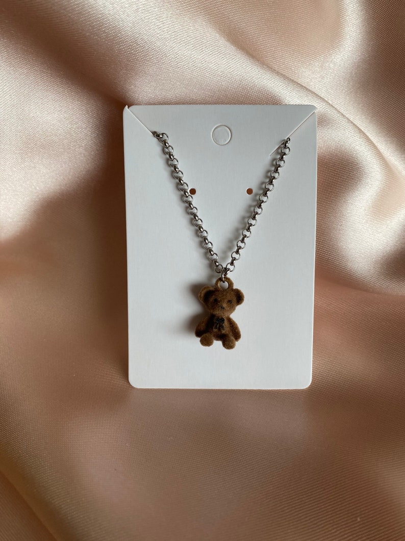 May include: A silver chain necklace with a brown teddy bear charm. The necklace is displayed on a white card.