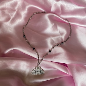 K&ouml;nnte beinhalten: Silberfarbene Halskette mit einem Anh&auml;nger in Form einer Motte mit filigranen Details und einem kleinen Mondakzent. Die Kette enth&auml;lt kleine schwarze Perlen. Pr&auml;sentiert auf rosa Satin.