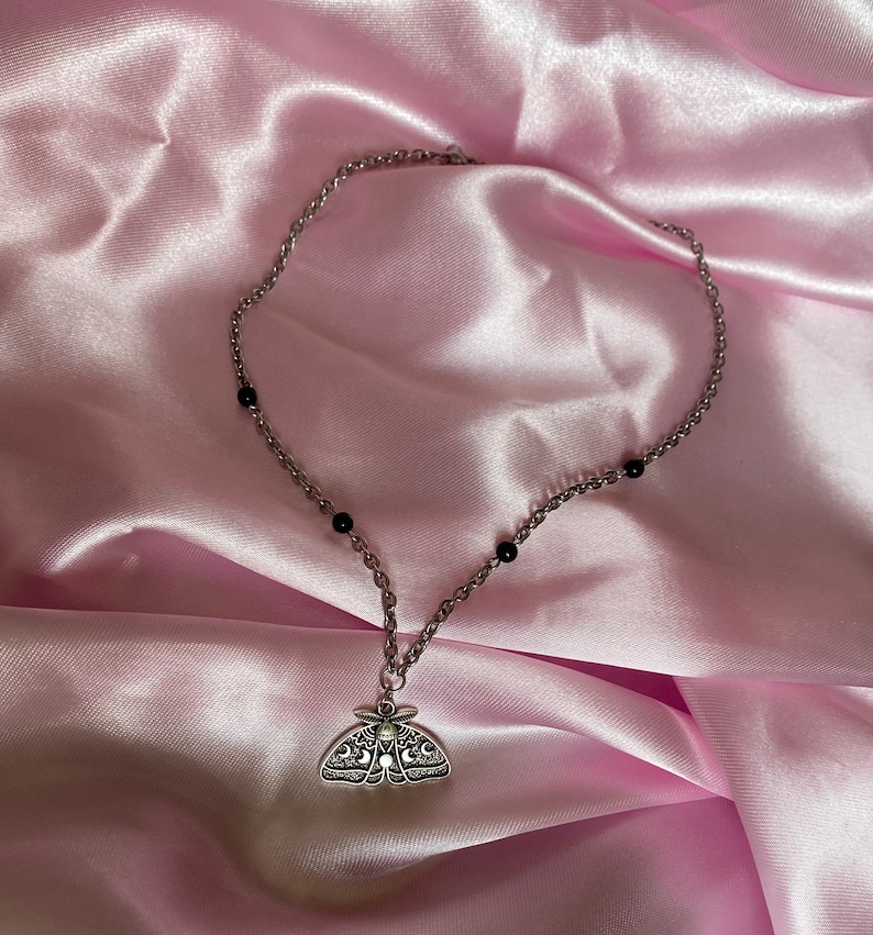 K&ouml;nnte beinhalten: Silberfarbene Halskette mit einem Anh&auml;nger in Form einer Motte mit filigranen Details und einem kleinen Mondakzent. Die Kette enth&auml;lt kleine schwarze Perlen. Pr&auml;sentiert auf rosa Satin.