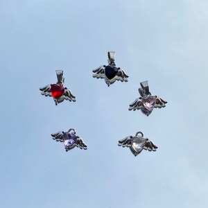 Op de afbeelding: Vijf zilveren hangers met vleugels en hartvormige edelstenen. De edelstenen zijn rood, zwart, roze, paars en helder.