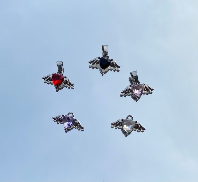 Op de afbeelding: Vijf zilveren hangers met vleugels en hartvormige edelstenen. De edelstenen zijn rood, zwart, roze, paars en helder.