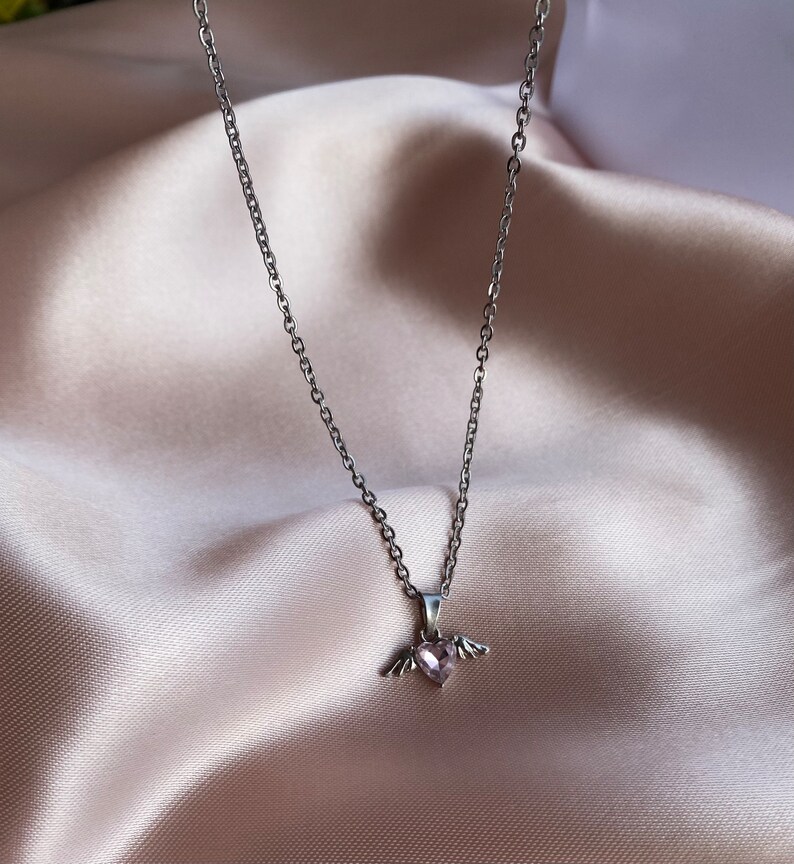 Op de afbeelding: Een zilveren ketting met een klein roze hartje met vleugels.