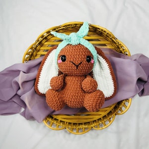 Puede incluir: Un conejito de crochet marrón con orejas blancas y una diadema de crochet azul claro. El conejito está sentado en una cesta tejida sobre un fondo de tela morado.
