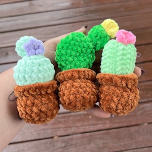 Puede incluir: Tres plantas de cactus de crochet en macetas marrones. Los cactus son verdes y tienen flores de diferentes colores en la parte superior. Las flores son moradas, amarillas y rosas.