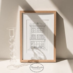 May include: A black and white line drawing of a building with a shop front, windows, and a sign that says "Meow's Studio". The drawing is framed in a light wood frame and is displayed on a white surface.