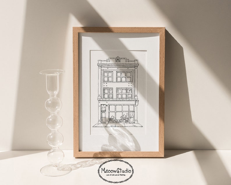 May include: A black and white line drawing of a building with a shop front, windows, and a sign that says "Meow's Studio". The drawing is framed in a light wood frame and is displayed on a white surface.