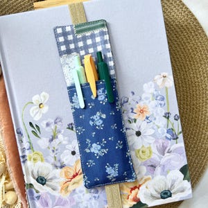 May include: A floral-covered journal with a blue pen holder containing three pens. The pen holder is attached to the journal with a beige elastic band. The journal cover features a watercolor floral design with white, purple, and orange flowers.