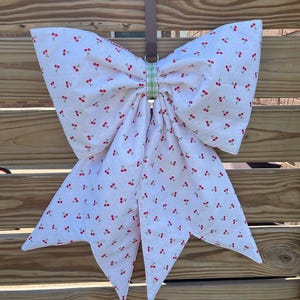 May include: A large fabric bow featuring a white background with a repeating pattern of small red cherries. The bow is held together by a green and white checkered fabric center loop and is suspended from a brown strap attached to a metal ring.