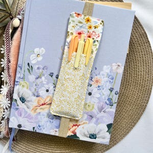May include: A light blue journal with floral illustrations, a gold ribbon bookmark, and a fabric pen holder filled with yellow and orange pens. The pen holder has a floral pattern on the front and is attached to the journal.