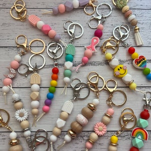 May include: A selection of keyrings with diverse, colourful bead designs. Keyrings feature gold and silver metal clasps, with beads in pastel, rainbow, and wooden tones. Some keyrings include decorative elements like flowers, rainbows, and smiley faces.