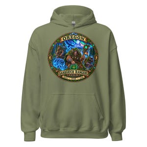 Puede incluir: Una sudadera con capucha de color verde oliva que presenta un gráfico circular en el pecho. El diseño muestra tres figuras de Sasquatch en un entorno forestal con el texto OREGON SASQUATCH RANGERS GUILD y PROTECT EXPLORE GUARD en el borde.