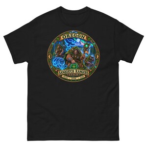May include: A black t-shirt featuring a circular graphic with the text OREGON SASQUATCH RANGERS GUILD PROTECT EXPLORE GUARD. The design depicts three figures in green cloaks in a forest setting with a glowing blue wolf and bird spirit.