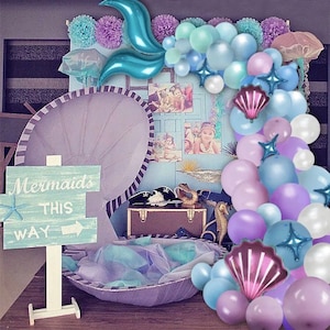 May include: A mermaid-themed party decoration with a large seashell backdrop, a sign that reads "Mermaids This Way", and a garland of blue, purple, and white balloons.