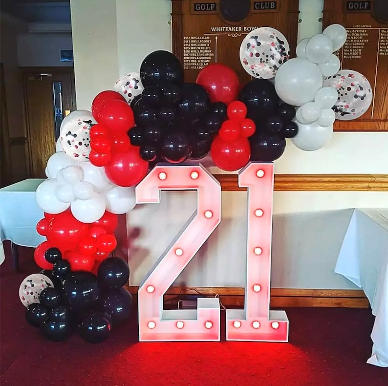 May include: A balloon arch made of red, white, and black balloons with confetti balloons. The arch is in front of two large light-up numbers that say "21".