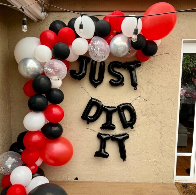 May include: A black balloon arch with red and white balloons, decorated with the words "JUST DID IT" in black balloons.