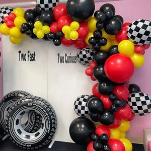 May include: A black, red, yellow, and white checkered balloon arch with the words "Two Fast" and "Two Curious" on a white backdrop. The arch is decorated with black, red, and yellow balloons. There are two black inflatable tires in the foreground.