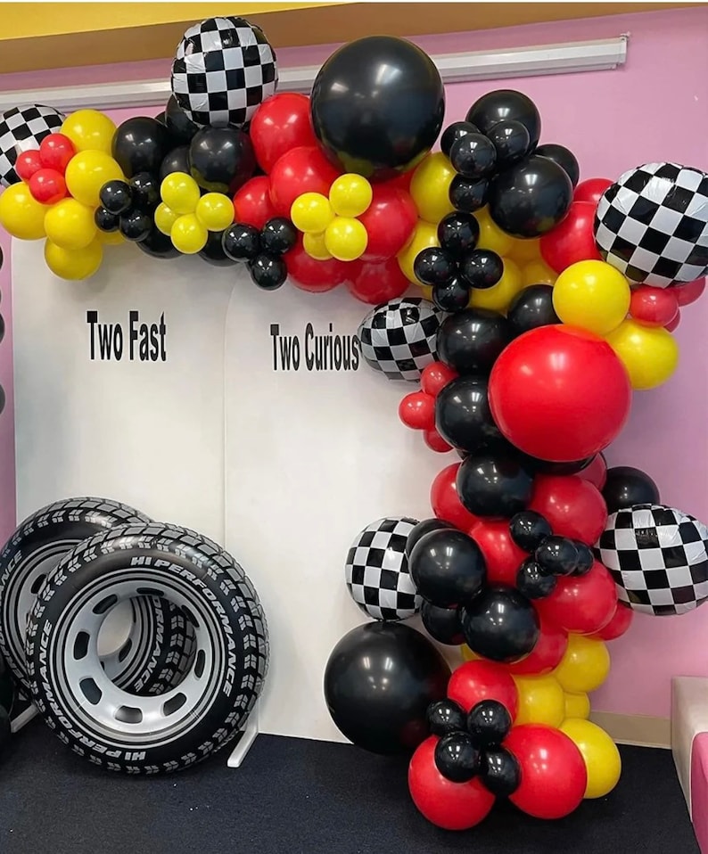 May include: A black, red, yellow, and white checkered balloon arch with the words "Two Fast" and "Two Curious" on a white backdrop. The arch is decorated with black, red, and yellow balloons. There are two black inflatable tires in the foreground.