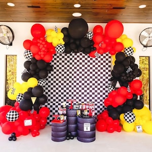 May include: A checkered backdrop with black and white squares is decorated with a balloon arch made of red, black, and yellow balloons. The arch is made of various sizes of balloons, including some large, round balloons. There are two black barrels with the text "Speed Limit 1" on one of them.