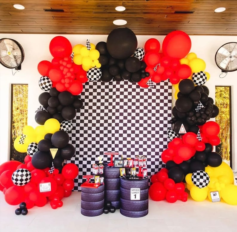 May include: A checkered backdrop with black and white squares is decorated with a balloon arch made of red, black, and yellow balloons. The arch is made of various sizes of balloons, including some large, round balloons. There are two black barrels with the text "Speed Limit 1" on one of them.