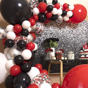 May include: A graduation party decoration with a balloon arch made of red, white, black, and silver balloons. The arch is decorated with confetti balloons. The background is a silver glitter backdrop. There is a table with cupcakes and other decorations.