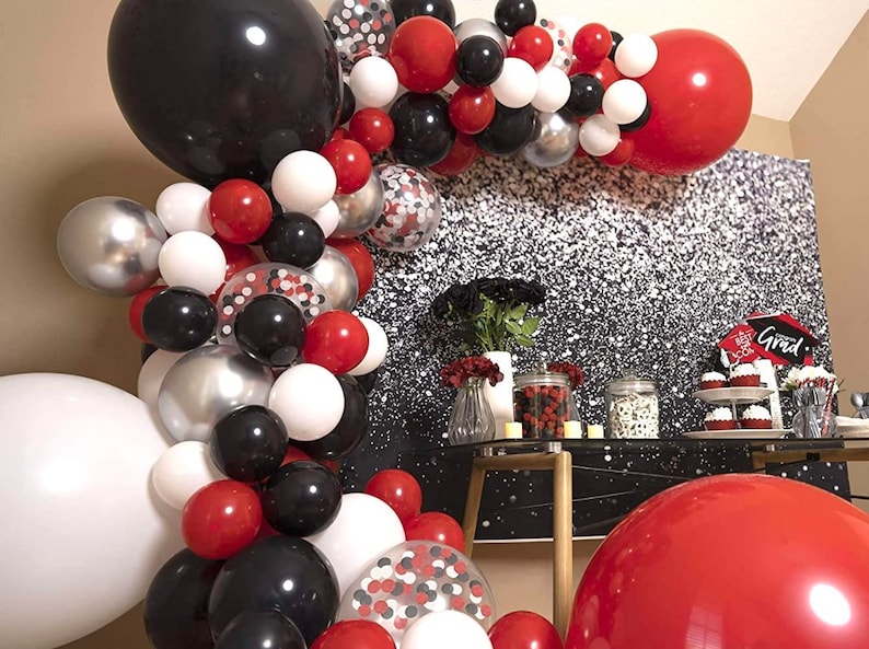 May include: A graduation party decoration with a balloon arch made of red, white, black, and silver balloons. The arch is decorated with confetti balloons. The background is a silver glitter backdrop. There is a table with cupcakes and other decorations.