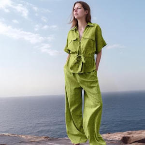 May include: A woman wearing a lime green linen outfit. The outfit includes a short-sleeved button-up shirt with two chest pockets and wide-leg pants. The shirt has a drawstring waist. The background shows a blue sky and ocean.