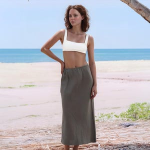 Pode incluir: Uma mulher vestindo um top branco de decote quadrado e uma saia longa verde-oliva. O look é apresentado em uma praia, com o mar e o céu ao fundo. A saia vai até os tornozelos.