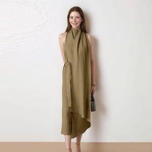 May include: A woman wearing a sleeveless olive green dress with a high neckline and a wrap-around tie at the waist. The dress has an asymmetrical hemline and is paired with a small black handbag.