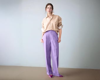 May include: A person wearing a tan button-down shirt with rolled-up sleeves and high-waisted, wide-leg lavender pants. The outfit is completed with magenta pointed-toe shoes. The background is a light blue wall.