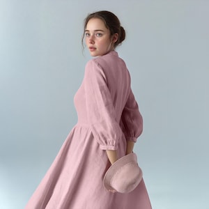 Op de afbeelding: Een vrouw die een roze linnen jurk draagt met een geplooide rok en pofmouwen. Ze houdt een roze hoed in haar rechterhand.
