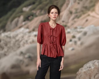 May include: A rust-colored blouse with short, puffed sleeves and a peplum hem. The top features a tie closure at the neckline and a gathered waist. Paired with black pants, the outfit is set against a blurred outdoor backdrop.