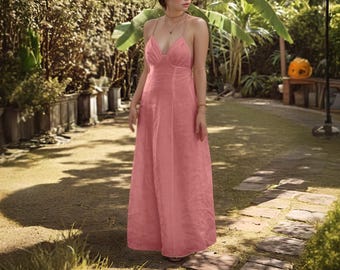 Puede incluir: Un vestido largo y fluido de color rosa con cuello en V y tirantes finos. El vestido tiene un corpiño ajustado y una falda larga hasta el suelo. La imagen está tomada al aire libre, con un jardín de fondo.