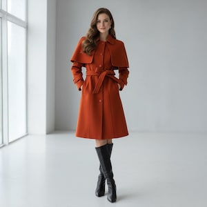 May include: A rust-colored coat with a cape-like overlay, button closures, and a matching belt. The coat features long sleeves and a flared skirt, paired with black knee-high boots. The woman is standing in a bright, minimalist space.