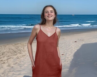 May include: A woman wearing a rust-colored sleeveless maxi dress with a V-neckline. She is standing on a sandy beach with the ocean in the background.