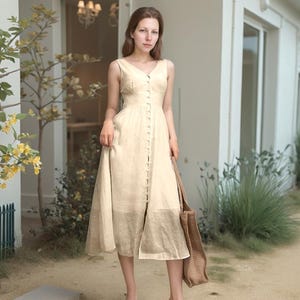 May include: A beige, button-down dress with a V-neck and a knee-length skirt. The dress is sleeveless and has a tailored bodice. A brown tote bag is held in the right hand. The background includes a white building and greenery.
