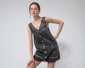 May include: A sleeveless, charcoal-coloured dress with a V-neck and white stitching. The dress features white buttons down the front and two front pockets. The model is wearing the dress.