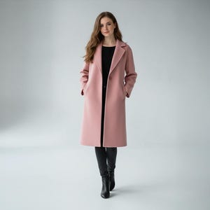 Puede incluir: Un abrigo largo de color rosa claro con solapas clásicas. El abrigo se lleva sobre una camiseta negra y pantalones negros. La modelo lleva botas negras. El abrigo tiene bolsillos y un corte entallado.