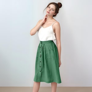 Puede incluir: Una falda verde con botones y un bolsillo en el lado derecho. La falda es de largo hasta la rodilla y tiene una cintura ajustada.