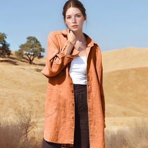 May include: A long, orange linen shirt worn over a white top and dark trousers. The shirt has a button-down front and a relaxed fit. The background shows a sunny landscape with rolling hills and trees.
