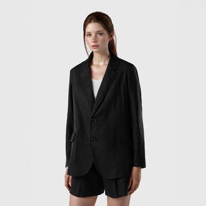 May include: A black blazer and matching shorts. The blazer has a notched lapel, two buttons, and a front pocket. The shorts are tailored with a pleated front. The outfit is worn over a white top. The model is standing in front of a white background.