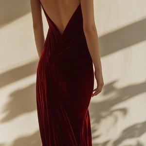 May include: A woman wearing a deep red velvet maxi dress with a low back and a slit in the skirt. The dress has a draped design and is fitted through the bodice.
