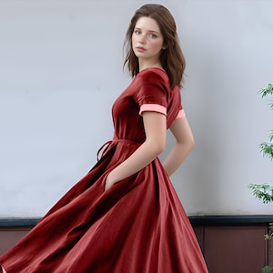 May include: A woman wearing a red linen dress with pink trim on the sleeves. The dress has a cinched waist and a full skirt.