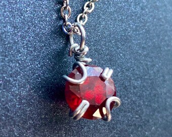 May include: A silver-toned necklace with a faceted, deep red gemstone pendant. The pendant is held in place by a wire cage design. The chain is delicate and the background is a dark, textured surface.