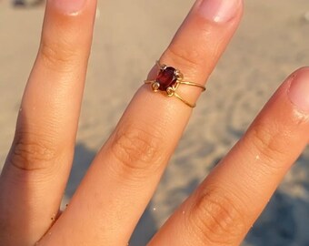 May include: A gold ring with a red gemstone set in the center. The ring is worn on a finger on a hand.
