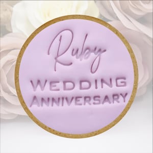 May include: A circular biscuit with a pale purple centre and a brown edge. The centre features the words "Ruby WEDDING ANNIVERSARY" embossed in a darker purple script and block letters.