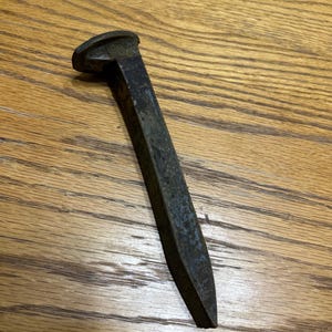 May include: A weathered, dark metal railway spike with a flat, rounded head and a tapered, rectangular shaft. The spike shows signs of age and use, with a slightly rusted appearance. The object is set against a wooden surface.