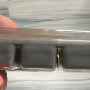 Puede incluir: Un recipiente de pl&aacute;stico transparente que contiene tres cubos de cera gris. Los cubos son rectangulares y parecen s&oacute;lidos. El recipiente lo sostiene una persona con las u&ntilde;as pintadas de naranja.