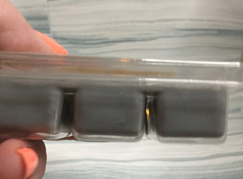 Puede incluir: Un recipiente de pl&aacute;stico transparente que contiene tres cubos de cera gris. Los cubos son rectangulares y parecen s&oacute;lidos. El recipiente lo sostiene una persona con las u&ntilde;as pintadas de naranja.